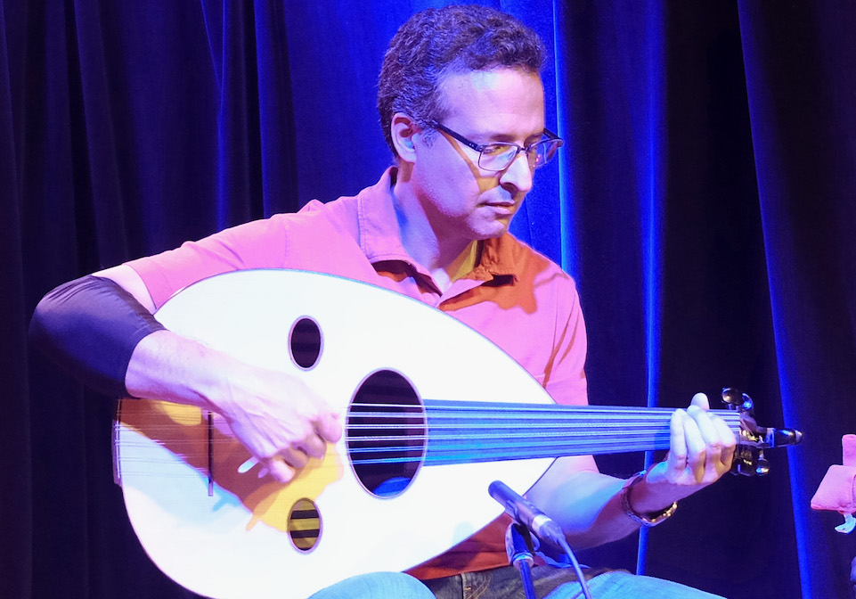 Khalil Moqadem, wearing eye-glasses and a light red shirt, plays the oud on stage in front of microphones.